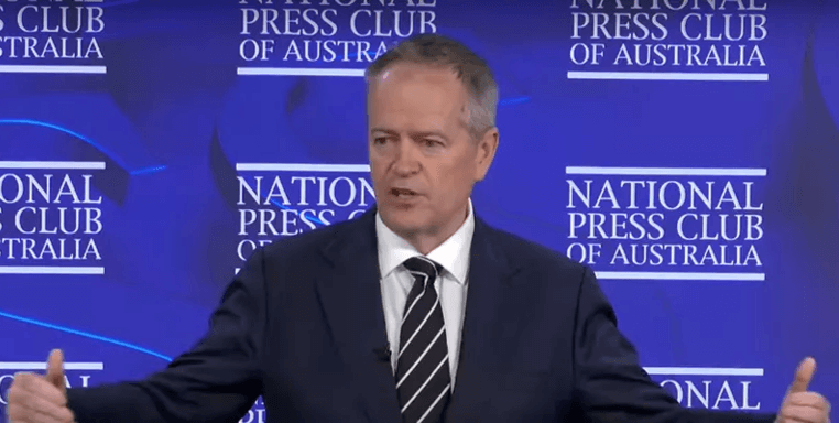Image of Minister Bill Shorten, a middle aged man with short greying hair wearing a black suit, white shirt and striped tie, speaking in front of a blue background with the National Press Club of Australia logo on it.